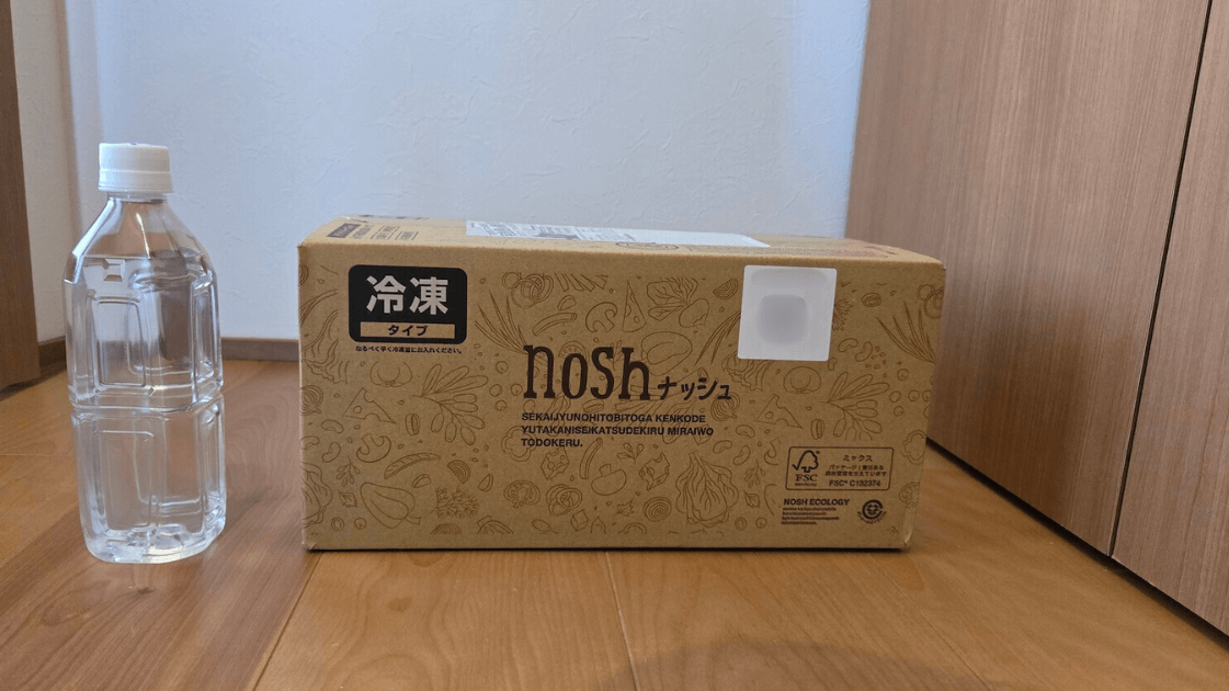 ナッシュの冷凍弁当が入った段ボールが玄関に届き、ペットボトルと並べてサイズ感がわかるように撮影された写真