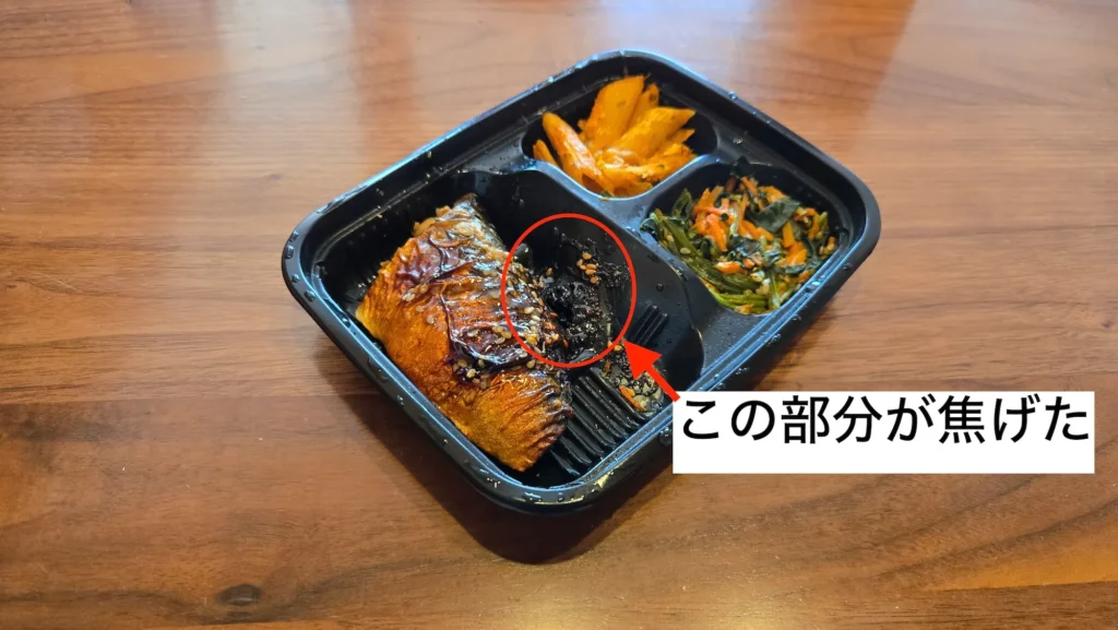 加熱後の弁当容器に焦げが残っているさばのごま照焼き。赤丸で焦げ部分を示した写真。