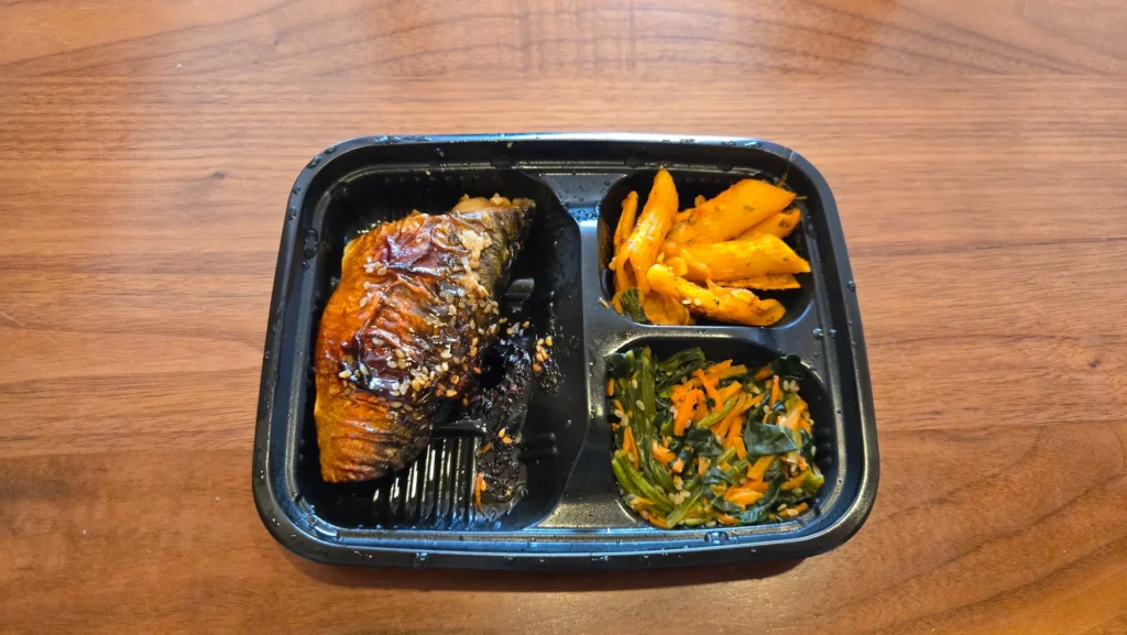 電子レンジで温めたさばのごま照焼き弁当。照りのあるサバに白ごまが散り、副菜にペンネとほうれん草のごま和えが添えられている。